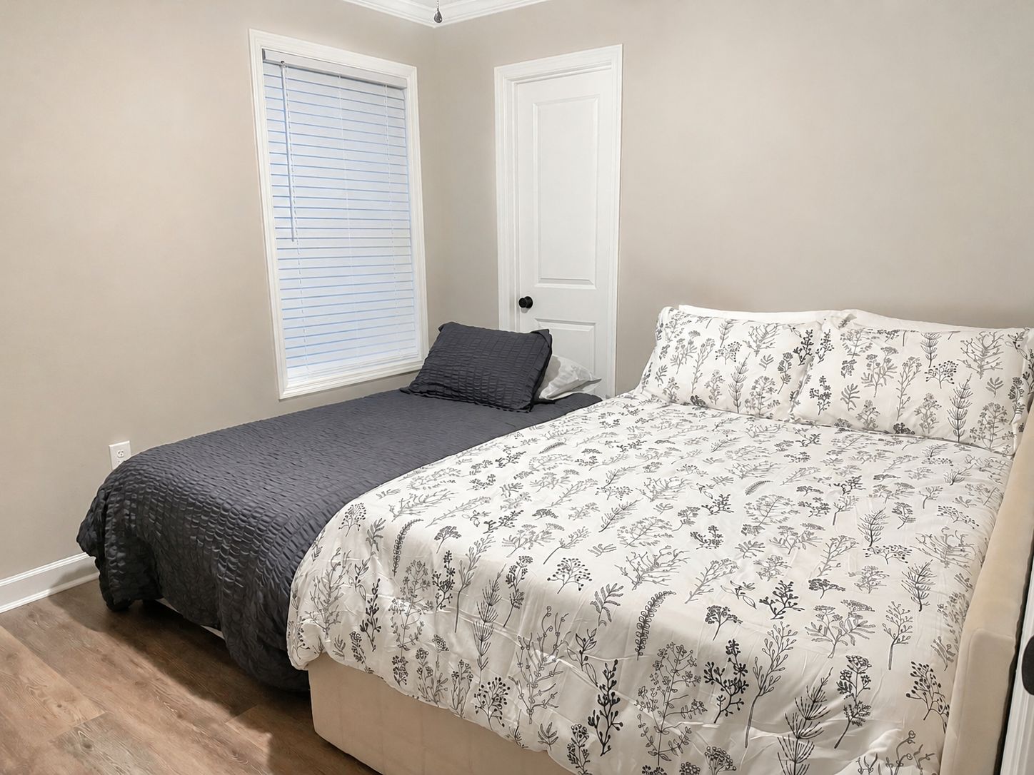 Une chambre à coucher avec un lit, une fenêtre avec des stores, une porte et un interrupteur sur le mur.