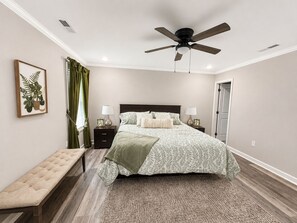 Un dormitorio con cama, mesitas de noche, un banco, ventilador de techo y planta enmarco.