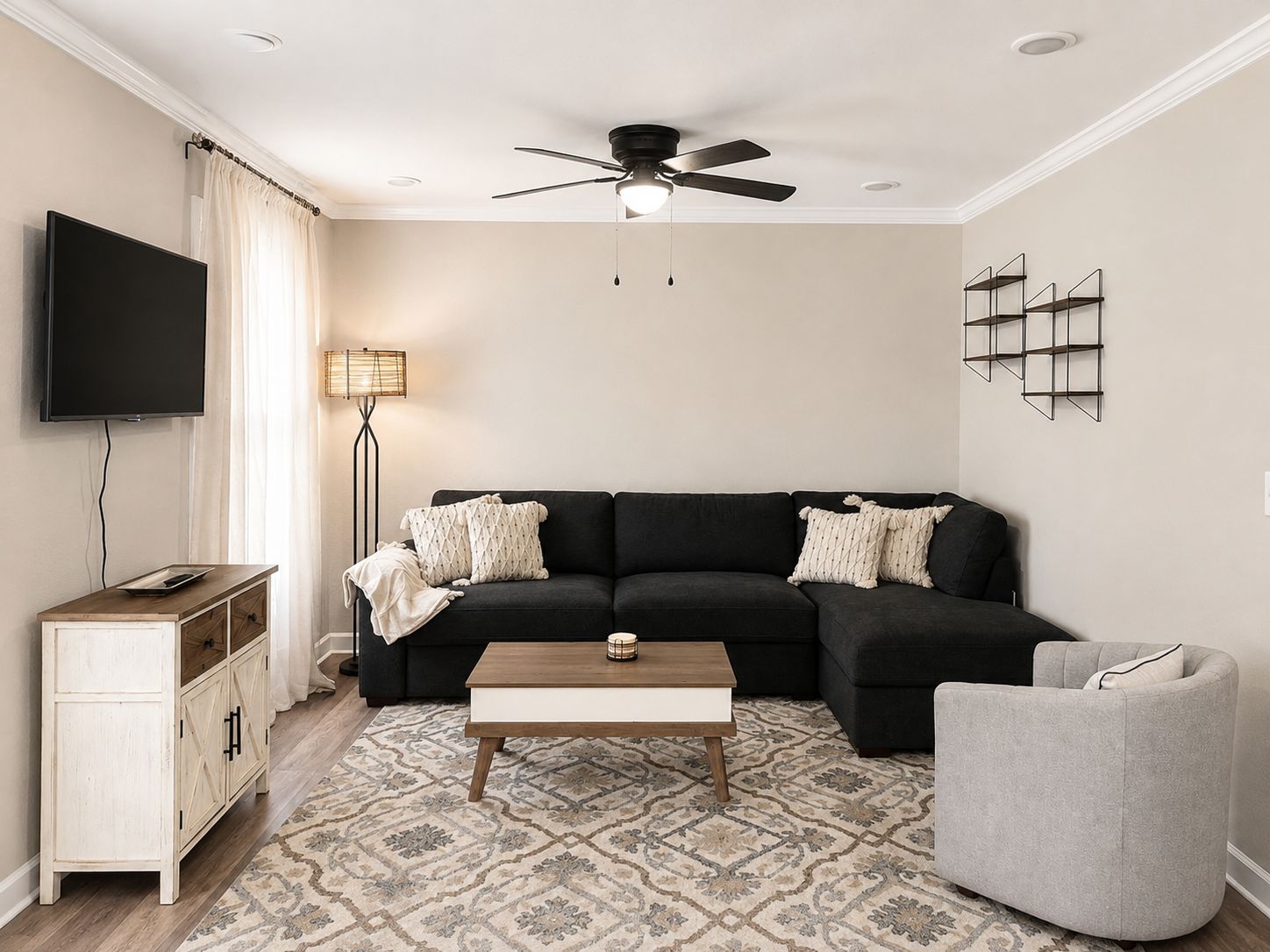 A living room with a sectional sofa, a coffee table, a TV, a floor lamp, a ceiling fan, and a bookshelf.