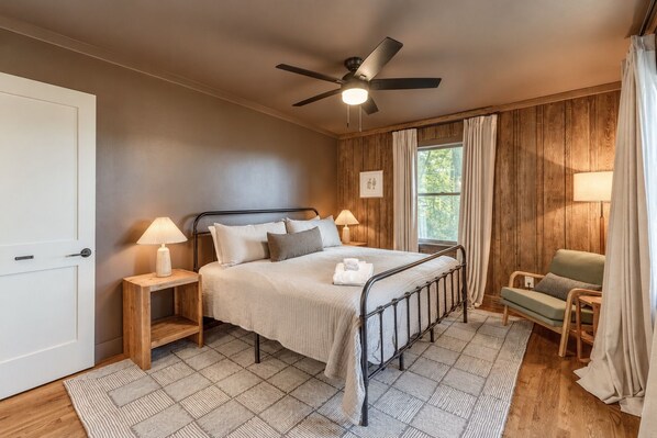 Ein Schlafzimmer mit Bett, Nachttischlampen, Deckenventilator, Fenster mit Vorhängen, Holzboden und Tür.