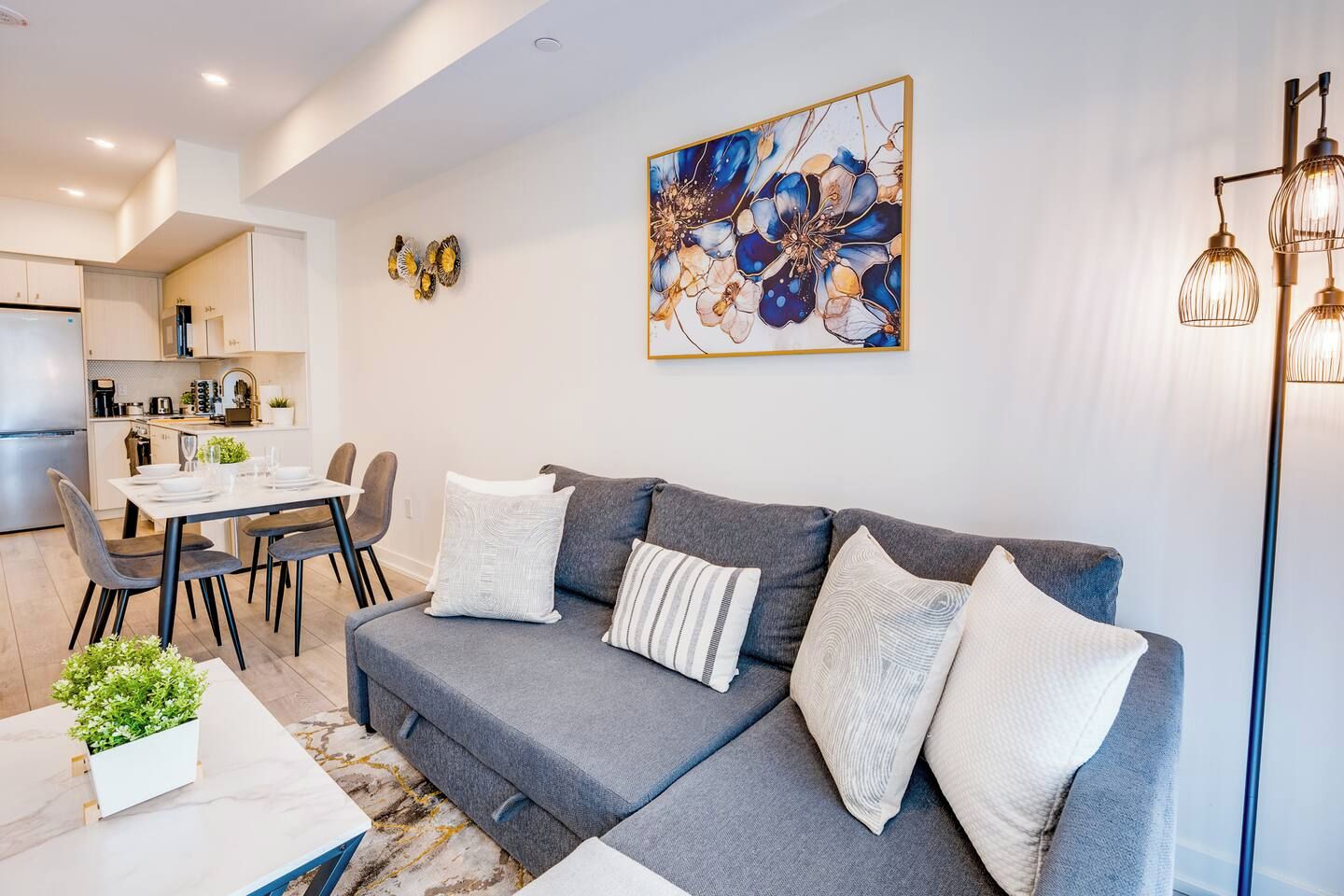Un salon moderne avec un canapé gris, des coussins blancs, un coin repas avec une table et des chaises blanches, et une cuisine en arrière-plan.