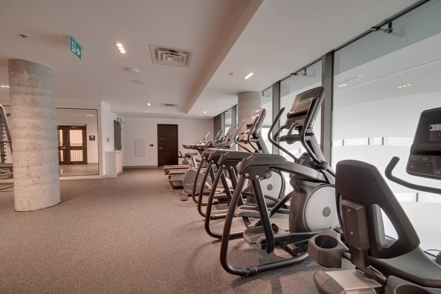 Une salle de sport avec des tapis de course alignés le long d’un mur, un pilier en béton et une porte à l’arrière-plan.