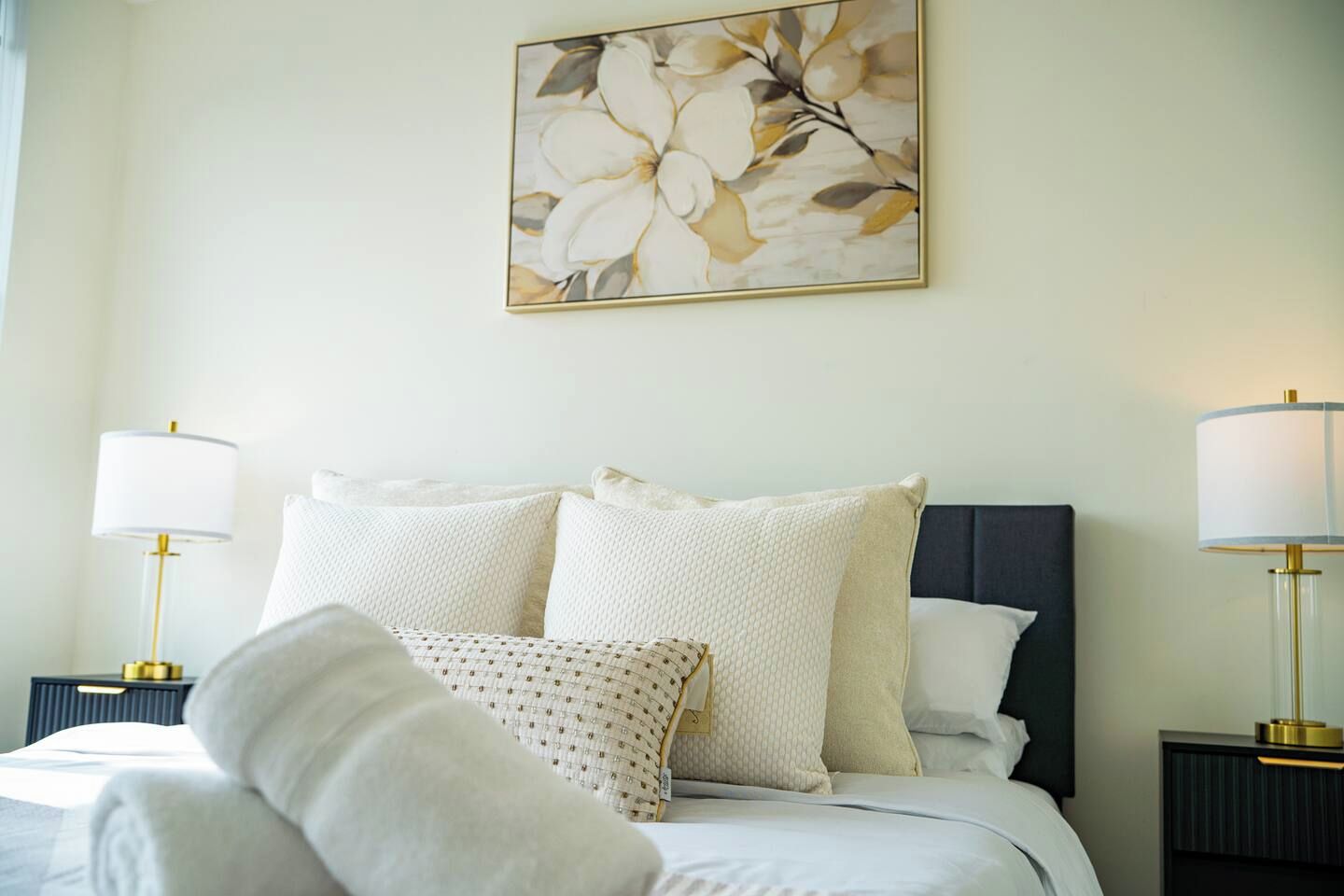 A neatly made bed with a white comforter and pillows, flanked by two bedside tables with lamps, in a room with a wall-mounted artwork above.