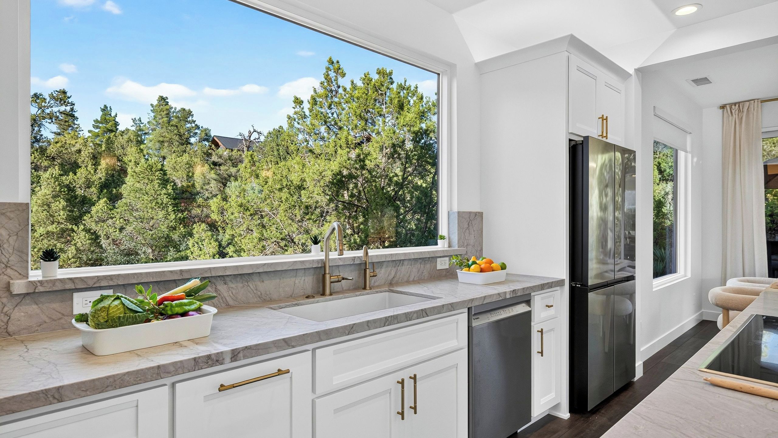 A modern kitchen with a large window, marble countertop, stainless steel appliances, and a view of trees.