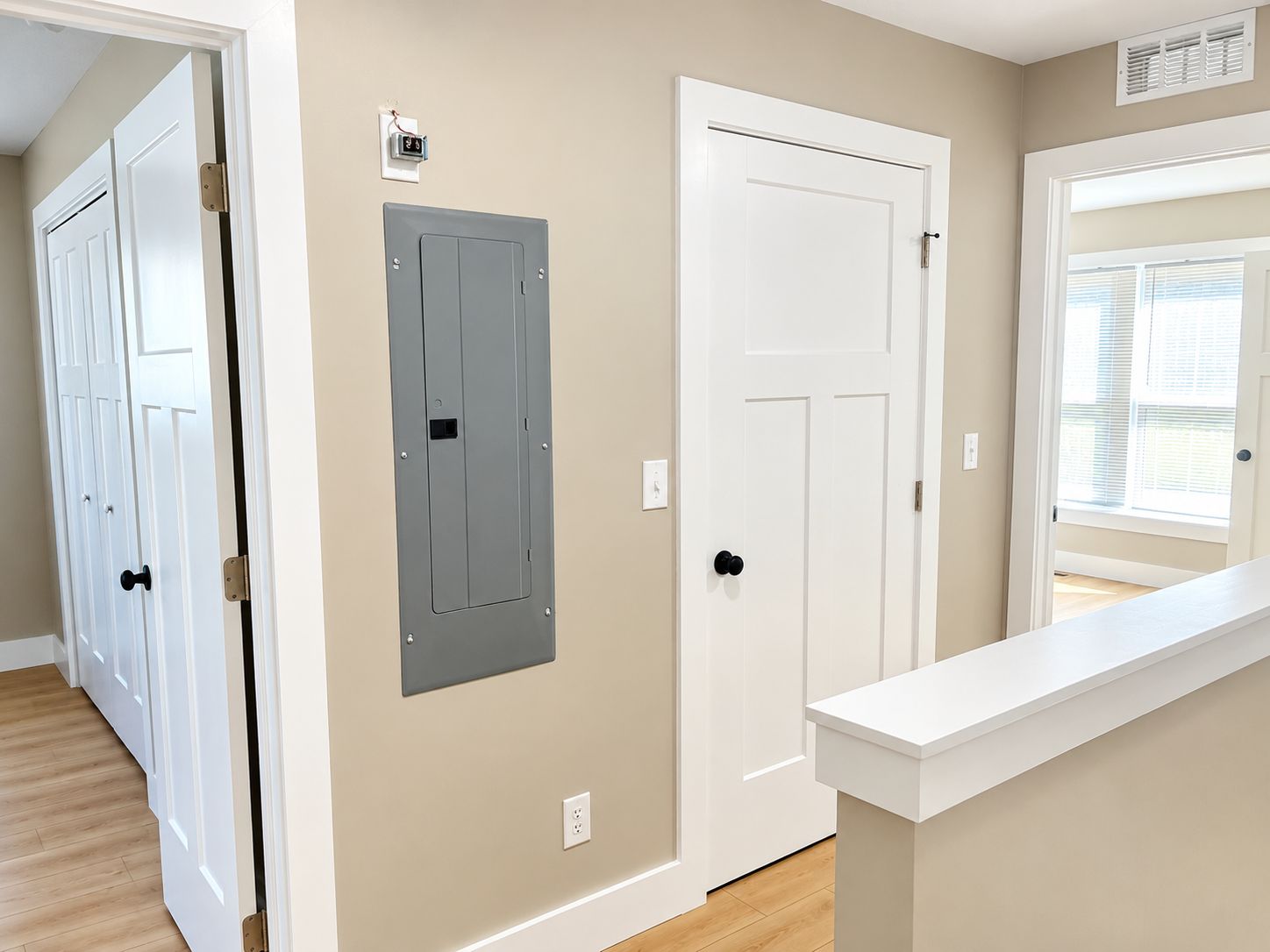 A hallway with a white door, a grey electrical panel, and a white wall with a window.