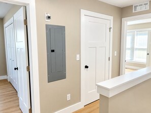 Un pasillo con una puerta blanca, un armario eléctrico gris y una pared blanca con un ventanal.