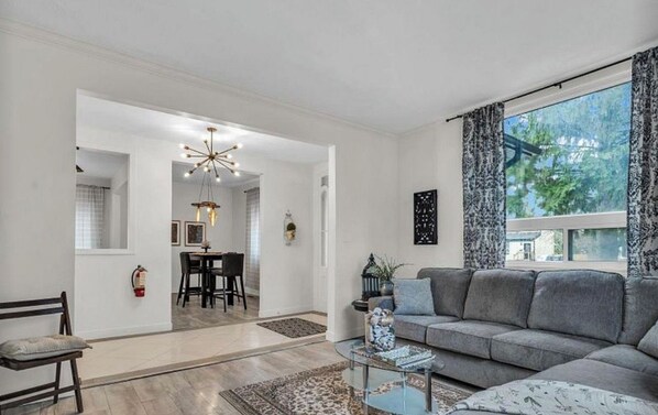 Een moderne woonkamer met een grijze bank, een glazen salontafel, en een eethoek met een kroonluchter.