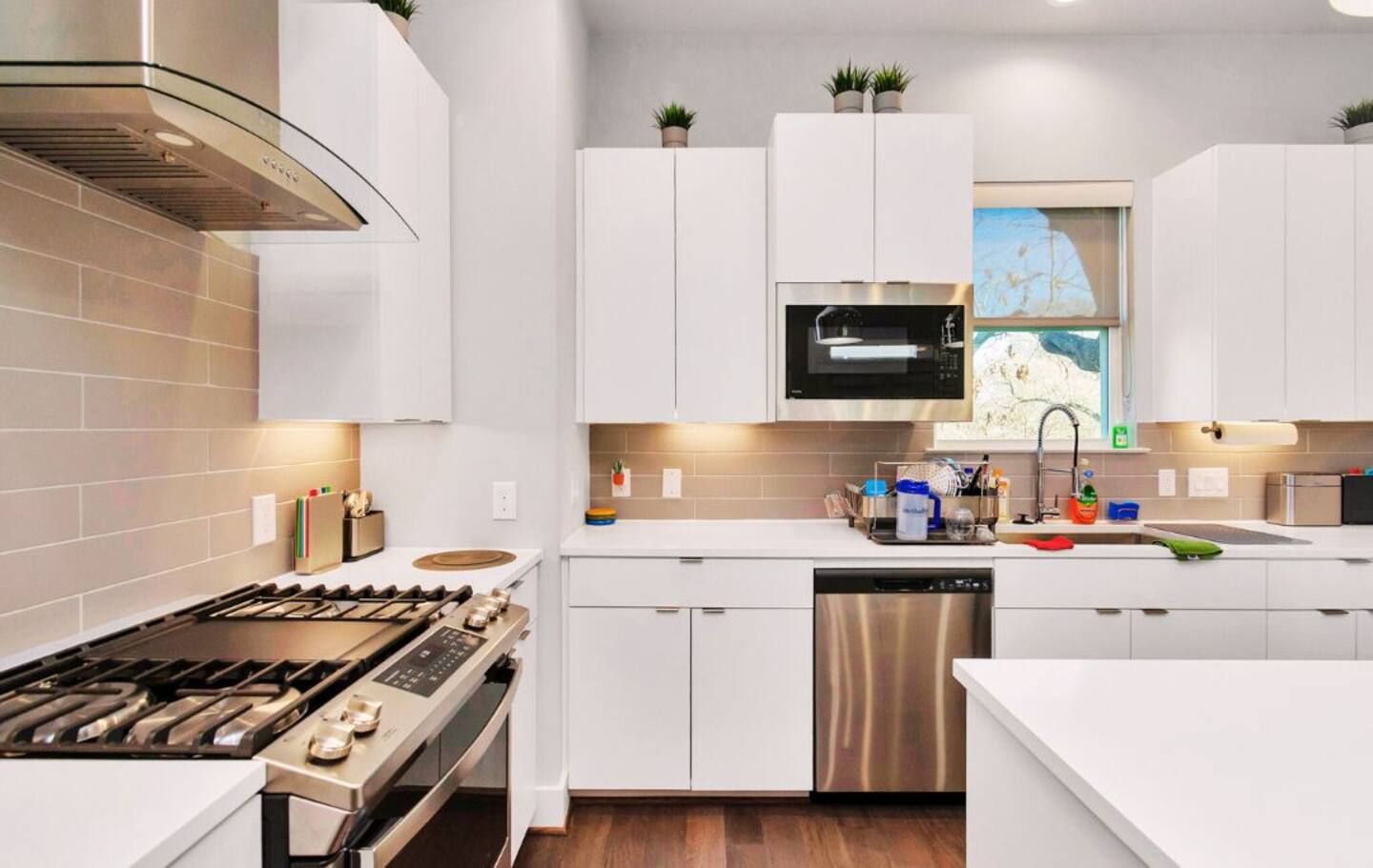 A modern kitchen with white cabinetry, a stainless steel range, a dishwasher, and a microwave. There are potted plants on the cabinets and a window with a view.