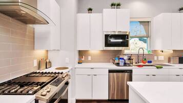 A modern kitchen with white cabinetry, a stainless steel range, a dishwasher, and a microwave. There are potted plants on the cabinets and a window with a view.