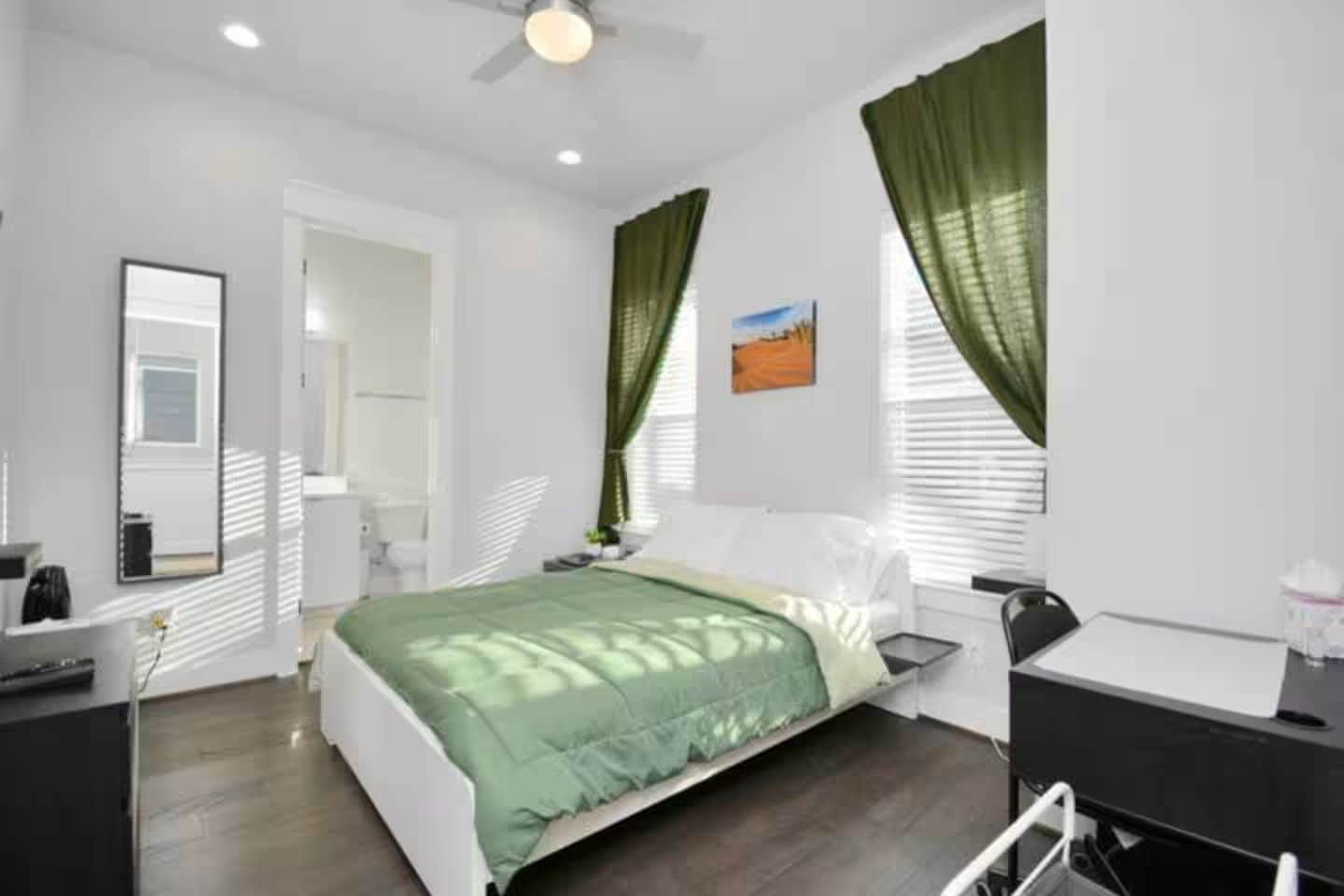 A bedroom with a bed, a desk, a chair, a mirror, and a window with curtains.