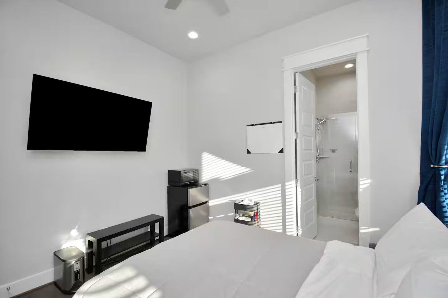 A modern hotel room with a bed, a flat-screen TV mounted on the wall, a small desk with a computer, and a shower area visible through an open door.