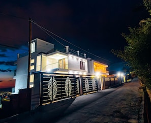 Una casa moderna de dos pisos con entrada protegida por un portón, un balcón visible y un camino de acceso a la vivienda.