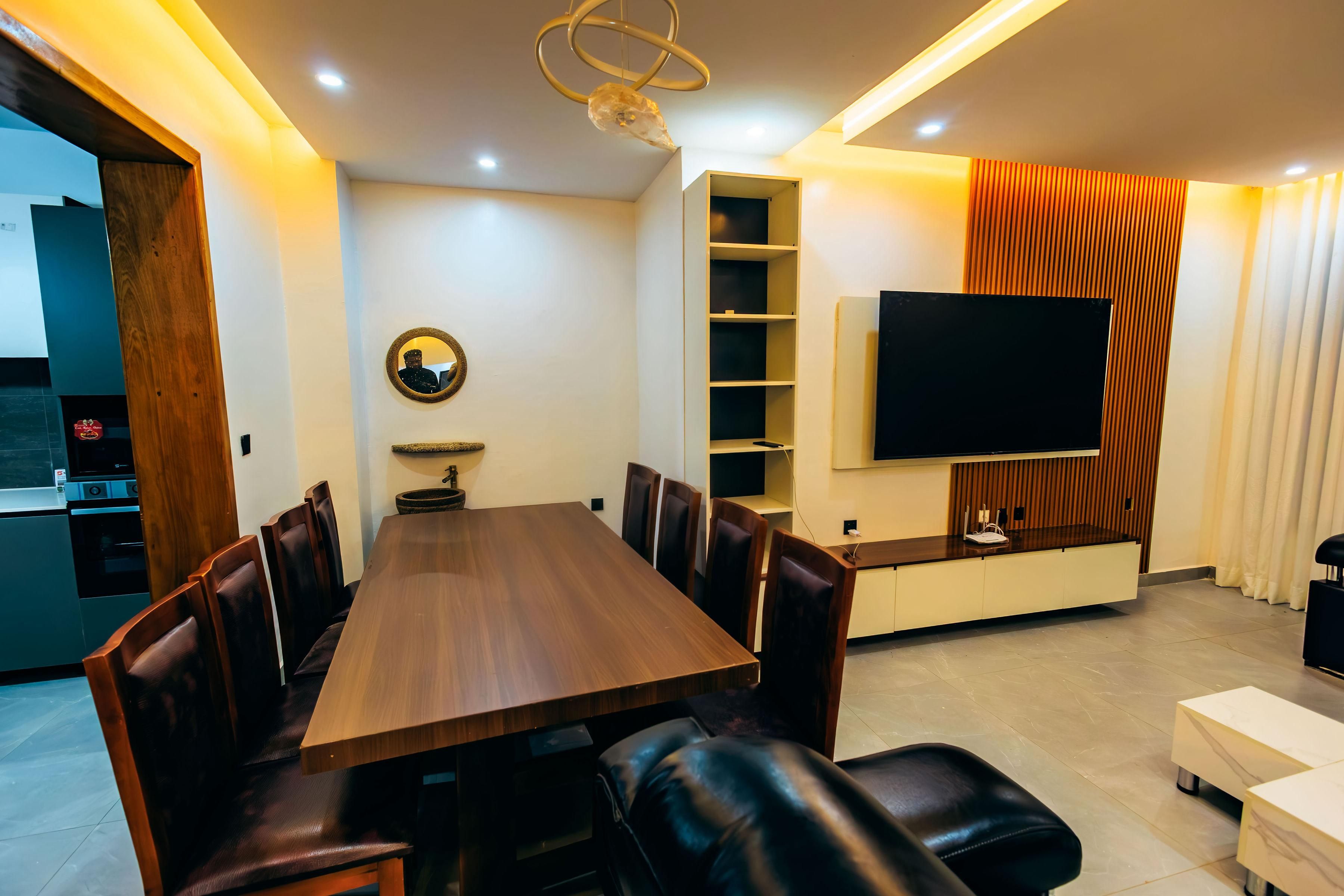 A modern dining area with a wooden table and leather chairs, a built-in TV, and a kitchen in the background.