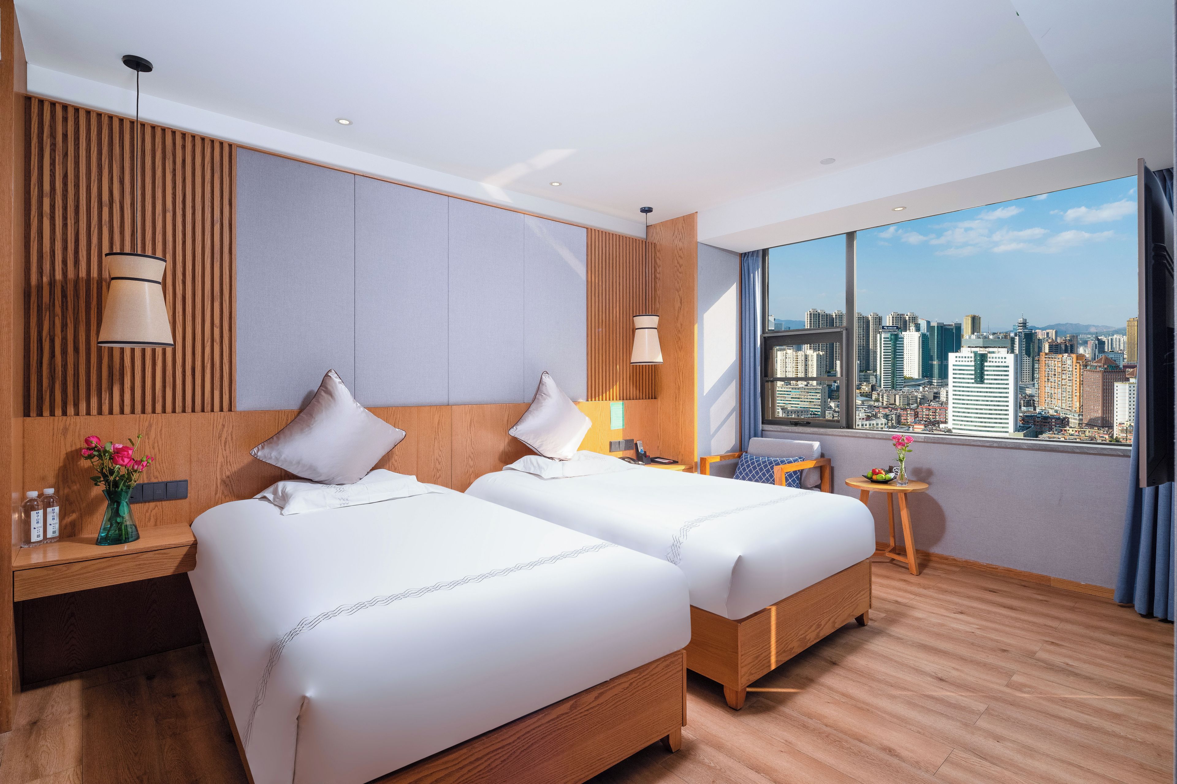 A hotel room with two beds, a city view, a wooden headboard, a small table with a vase of flowers, and a nightstand with a lamp.