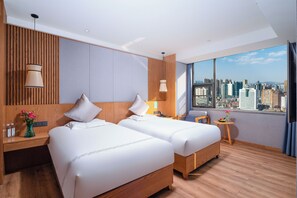 Habitación de hotel con dos camas, vista a la ciudad, cabecera de madera, una mesita con un florero y una mesita de noche con una lámpara.