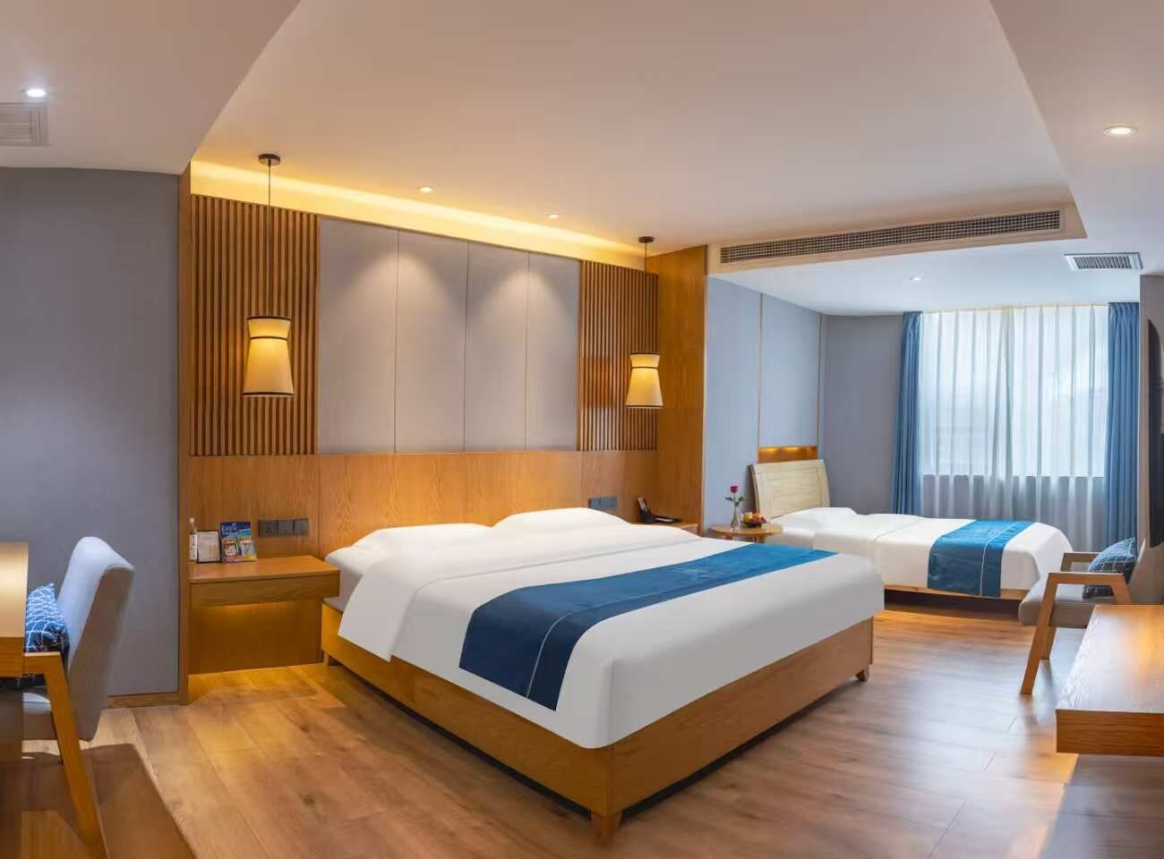 A hotel room with a large bed, a smaller bed, a desk, and a chair. There is a window with curtains, a wall-mounted lamp, and a ceiling vent.