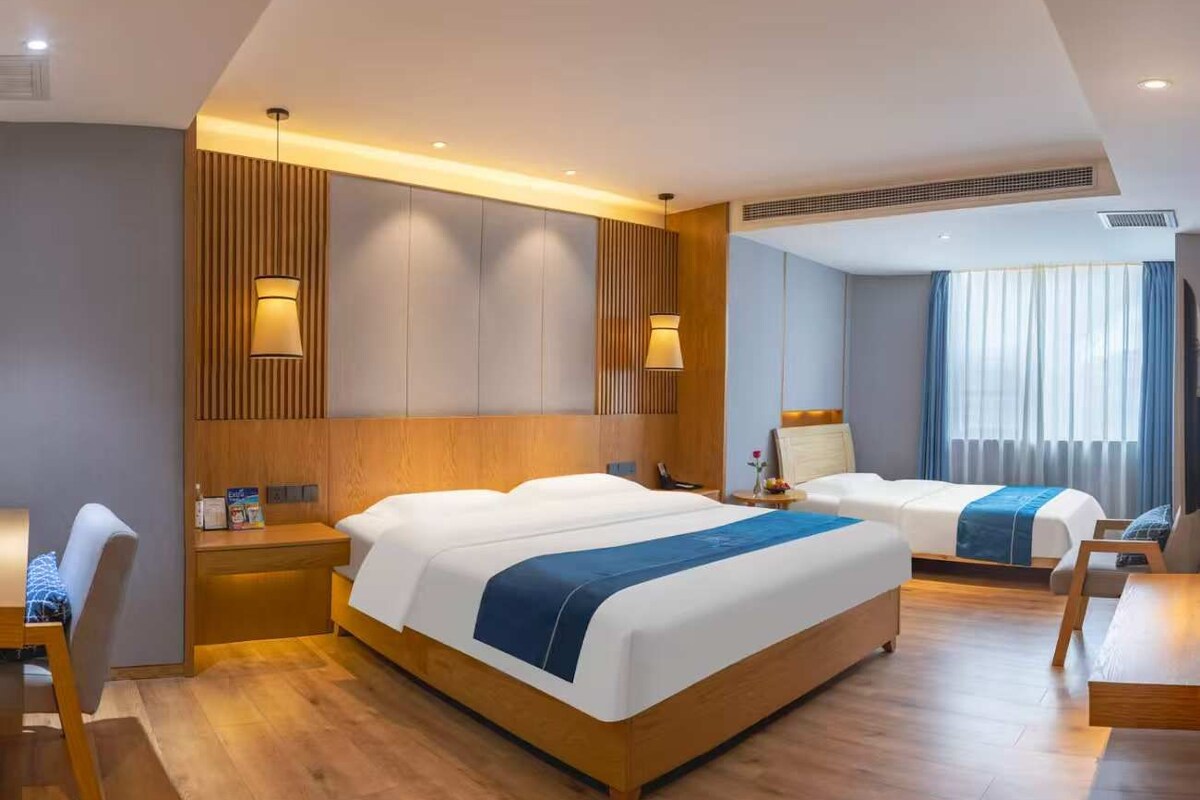 A hotel room with a large bed, a smaller bed, a desk, and a chair. There is a window with curtains, a wall-mounted lamp, and a ceiling vent.