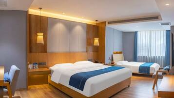 A hotel room with a large bed, a smaller bed, a desk, and a chair. There is a window with curtains, a wall-mounted lamp, and a ceiling vent.