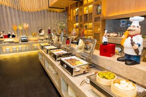 Zona de bufé con diferentes estaciones de comida, incluyendo una parrilla caliente, un bol de frutas y una selección de pasteles.