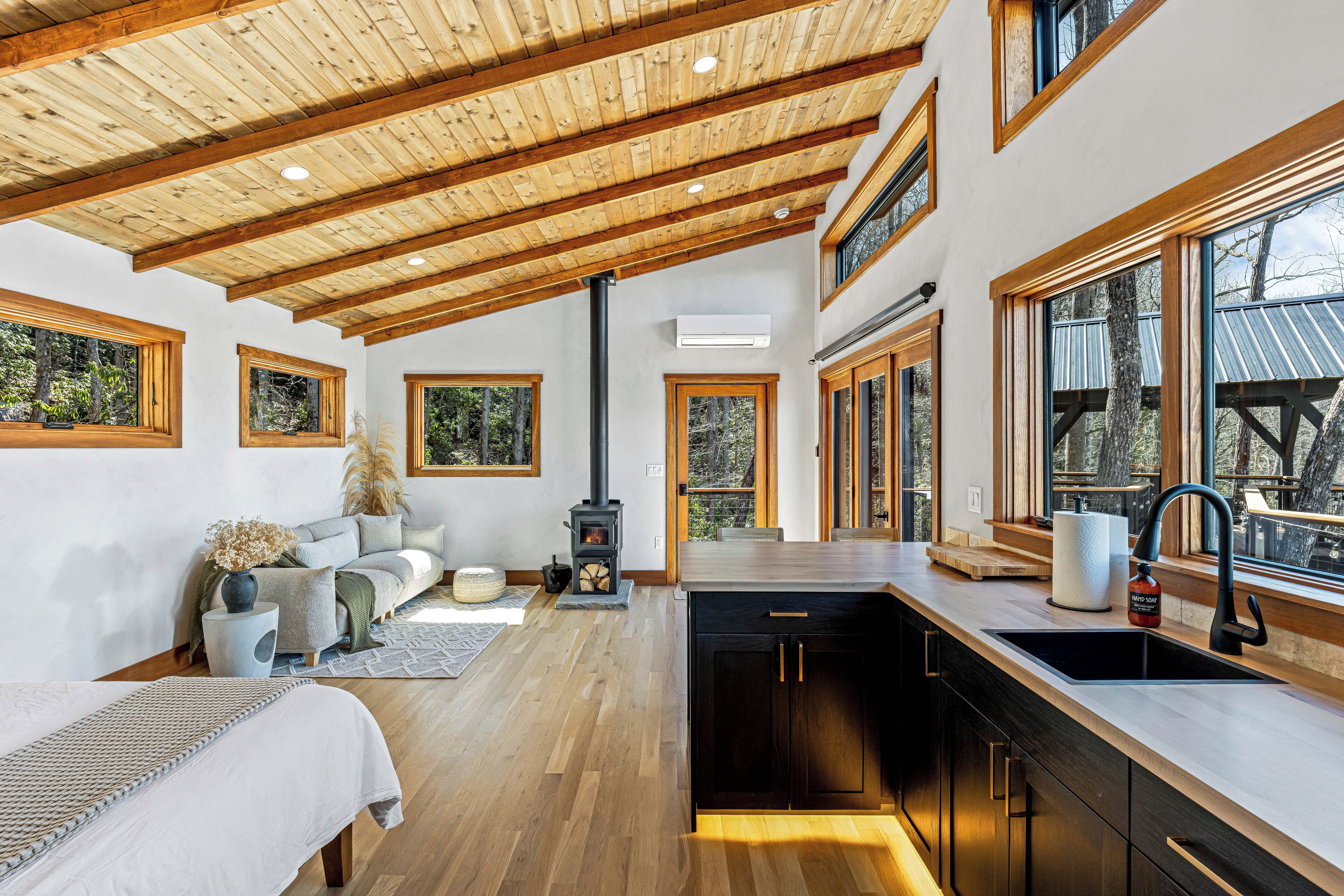 A modern interior with a wooden ceiling, large windows, a kitchen with a black stove, and a seating area with a sofa and decorative plants.