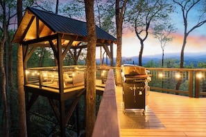 A wooden deck with a grill and seating area, surrounded by trees and overlooking a mountainous landscape. - Outdoor movies w Hot Tub, Fire Pit, Mtn Views (Penrose)