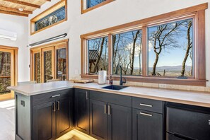 A modern kitchen with dark cabinetry, a large island with a sink, and large windows offering a view of trees and mountains. - Outdoor movies w Hot Tub, Fire Pit, Mtn Views (Penrose)