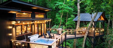 A cabin with a hot tub, deck, and outdoor seating area surrounded by forest.