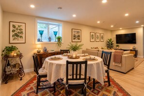A dining area with a round table, chairs, and a rug. There is a window with blinds, a sofa, and a television mounted on the wall. - Central Calgary 2 bedroom 1 bath Basement Suite with kitchen and living room. (Calgary)