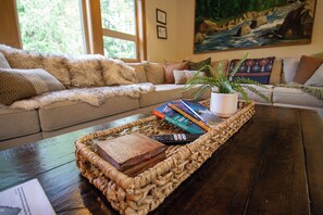 A living room with a sofa, a coffee table with a woven tray, books, a remote control, and a potted plant. - Roaring River Lodge - Riverfront, Fire Pit, EV (Rhododendron)
