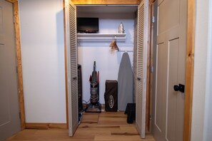 Passadís estret amb un terra de fusta, un televisor fixat a la paret, un aspirador i una taula de planxa plegable.