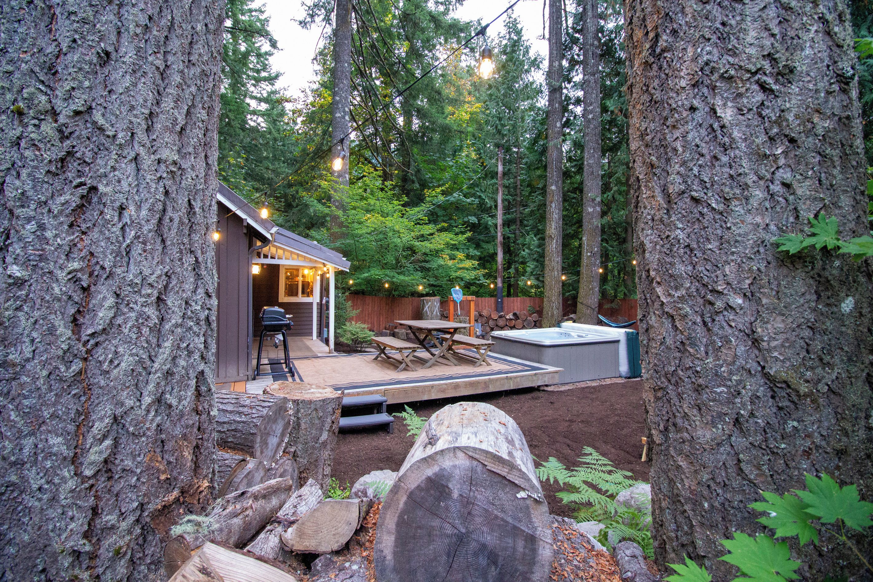 A cabin with a deck, outdoor seating, and a hot tub in a forested area.