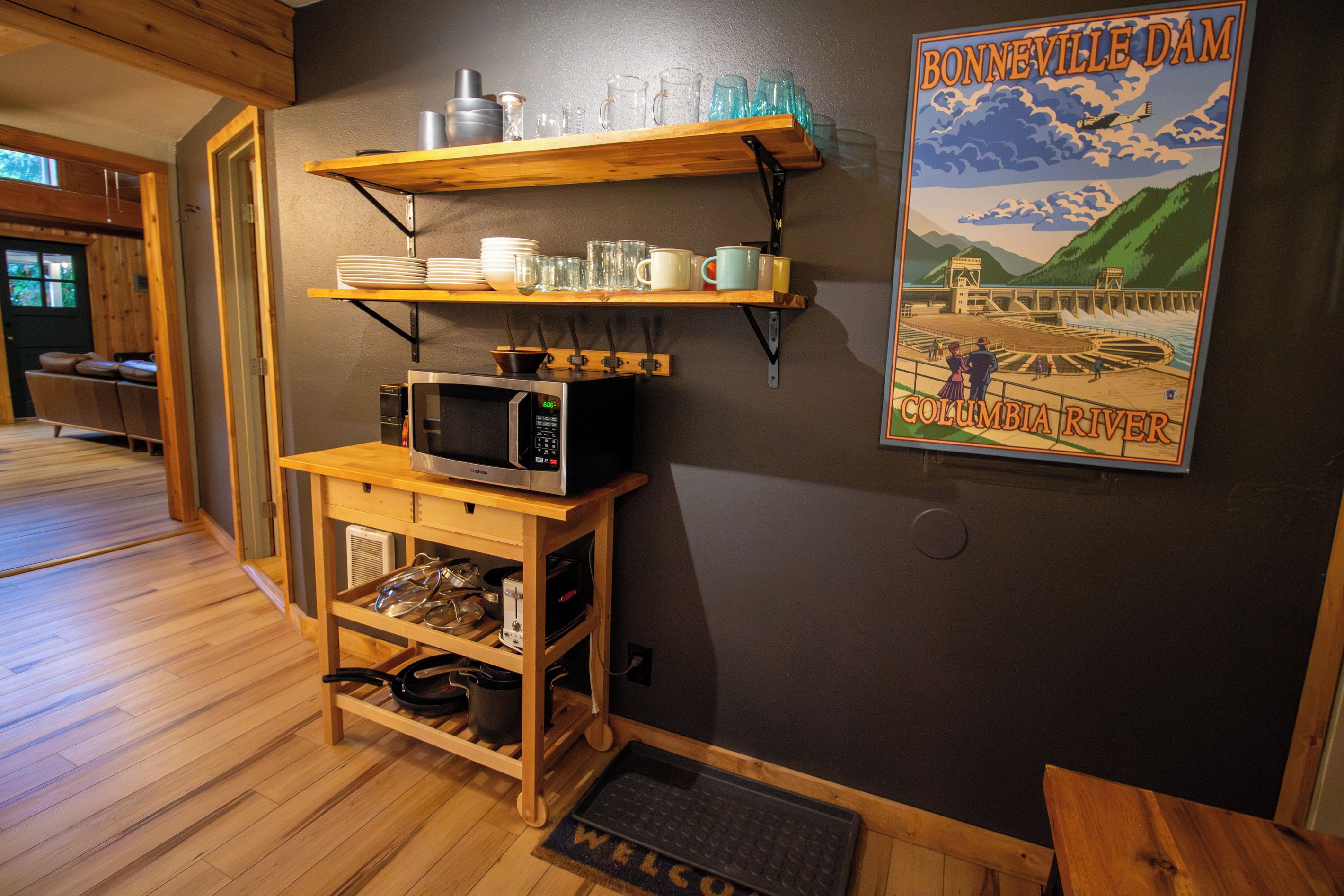 A kitchen with wooden shelves holding dishes and glasses, a microwave, and a poster of Bonneville Dam.