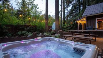 A hot tub with a wooden deck and picnic table, surrounded by trees and a wooden fence.