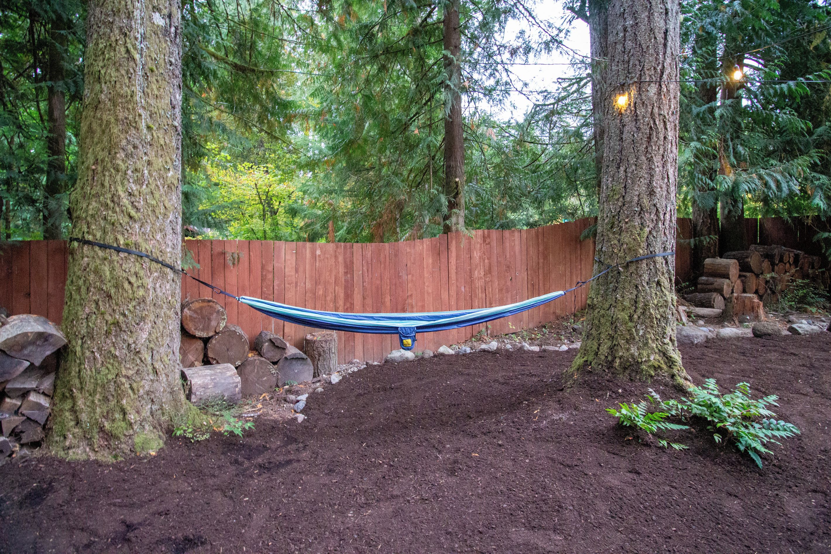 Un hamac tendu entre des arbres dans une zone boisée, avec une clôture en bois et une pile de bûches.