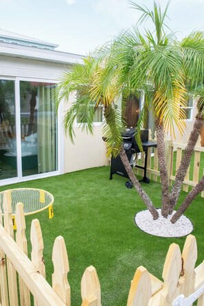 A backyard with artificial grass, a wooden fence, a barbecue grill, and a palm tree.