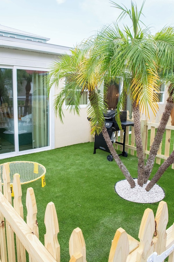 Un patio trasero con césped artificial, cerca de madera, una parrilla de barbacoa y una palmera.