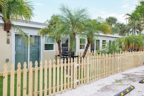 A row of single-story buildings with a wooden picket fence, surrounded by palm trees and a gravel driveway.