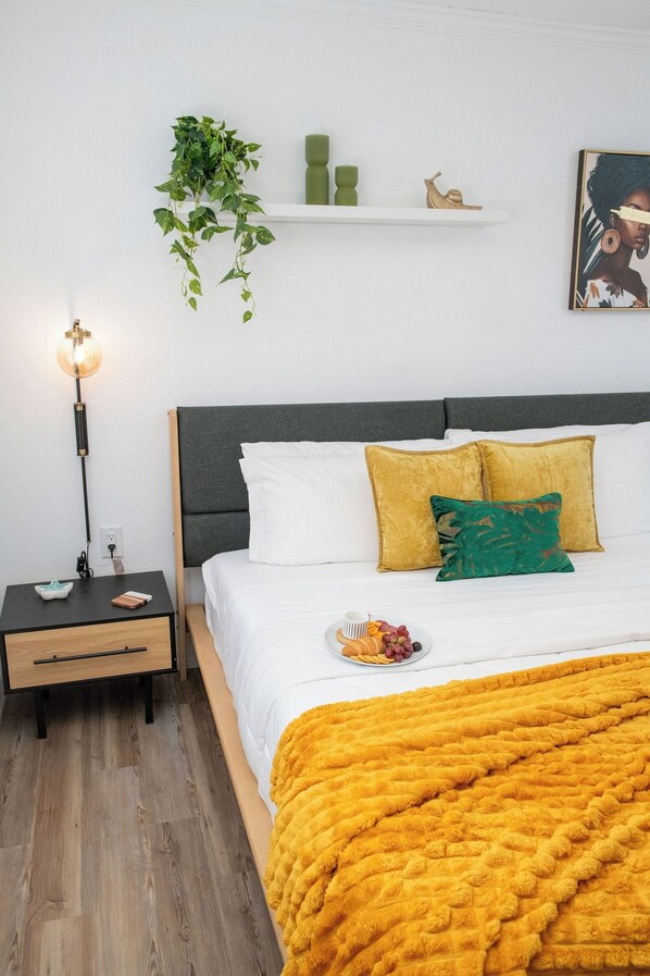 A bedroom with a bed, bedside table, lamp, wall-mounted shelf, and a painting.