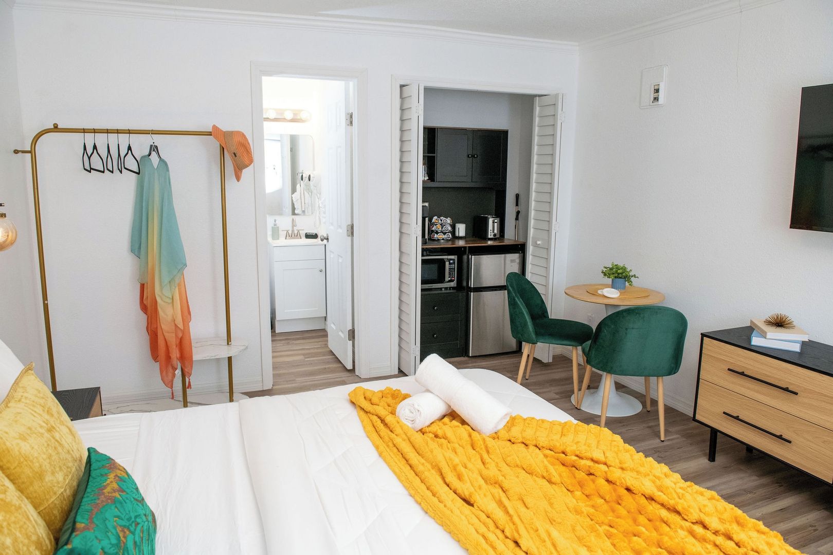 A hotel room with a bed, a desk, a chair, a TV, and a kitchenette.
