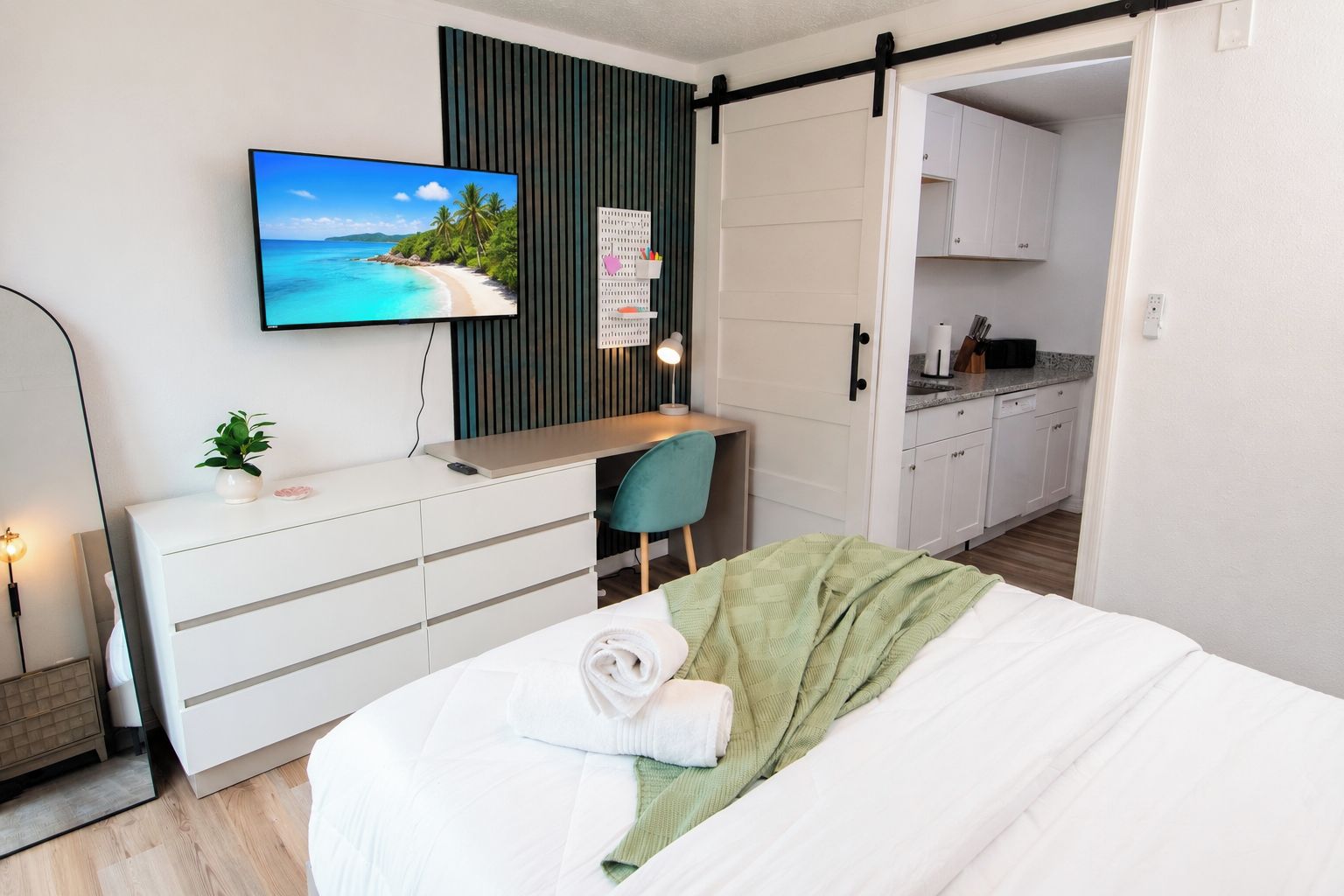 A bedroom with a bed, a desk with a computer, a TV, and a view of a kitchen.