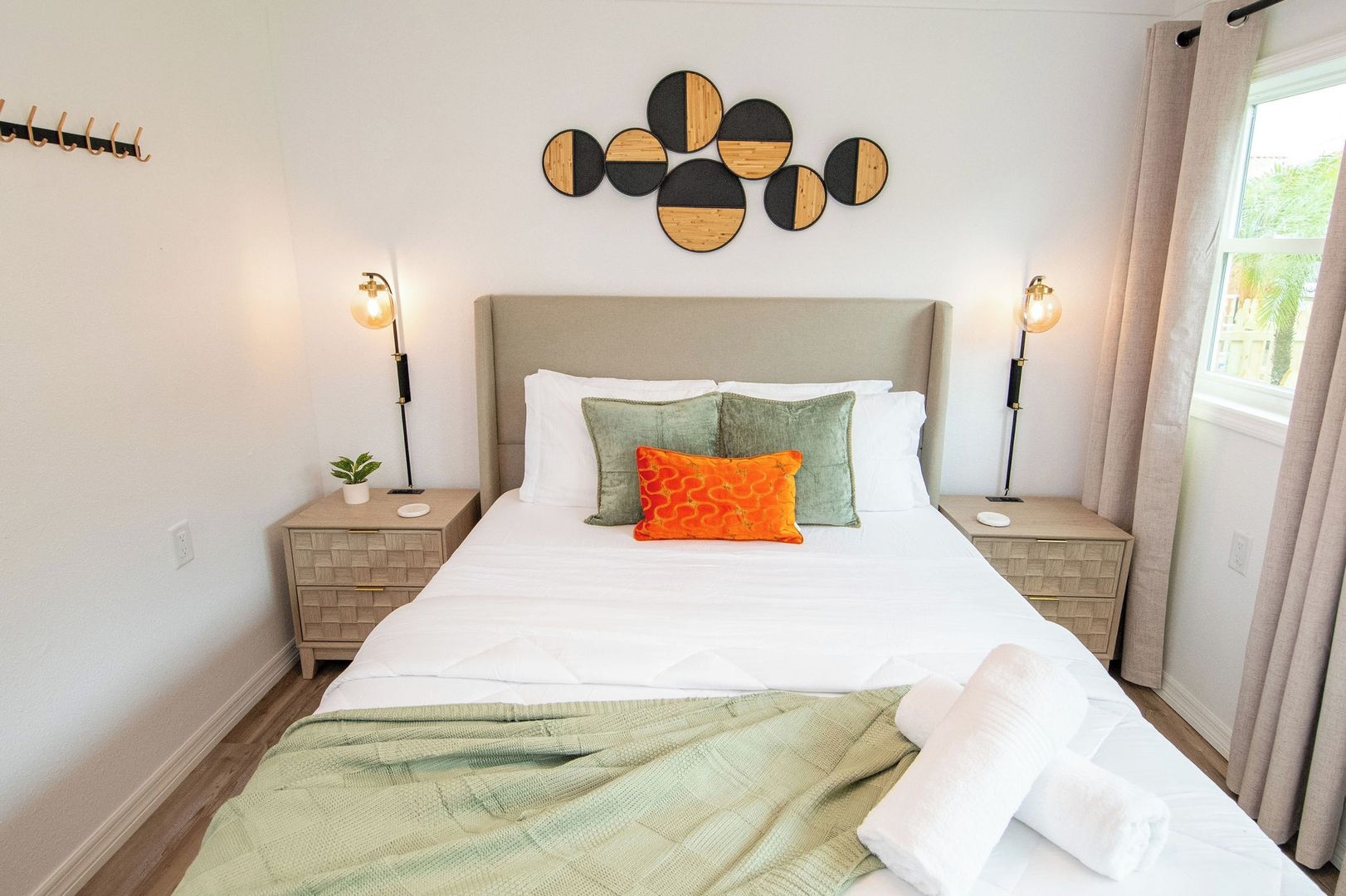 Ein ordentlich bezogenes Bett mit einer grünen Decke und einem orangefarbenen Kopfkissen, zwei Nachttische mit Lampen sowie ein Wandbild über dem Bett.