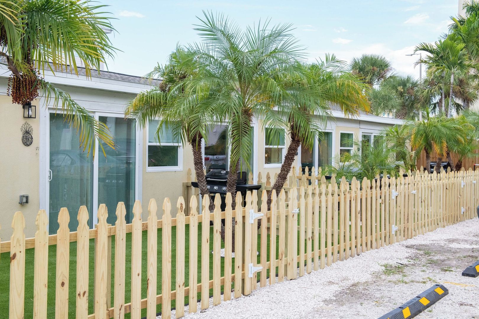 A row of single-story buildings with a wooden picket fence, surrounded by palm trees and a gravel driveway.