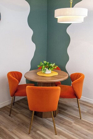 Un coin repas avec une table ronde en bois, quatre chaises orange, une plante en pot et un luminaire suspendu moderne.