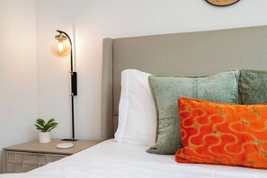 A neatly made bed with a grey headboard, a bedside lamp, a small potted plant, and two pillows with a green and orange pattern.