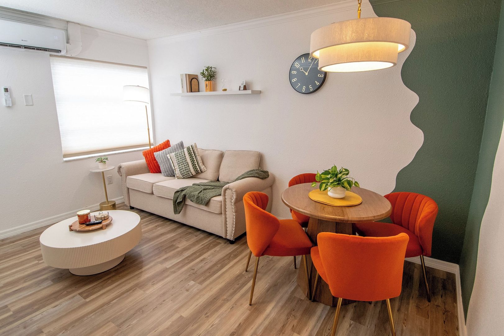 A modern living room with a sofa, a dining table with chairs, a round coffee table, a wall clock, and a hanging light fixture.