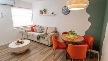 A modern living room with a sofa, a dining table with chairs, a round coffee table, a wall clock, and a hanging light fixture.