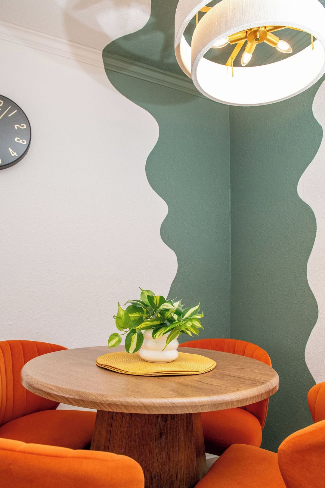 Uma mesa de madeira redonda com uma planta em vaso, rodeada por cadeiras laranja, num quarto com parede de destaque verde e luminária moderna.