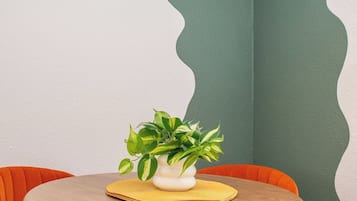 Uma mesa de madeira redonda com uma planta em vaso, rodeada por cadeiras laranja, num quarto com parede de destaque verde e luminária moderna.