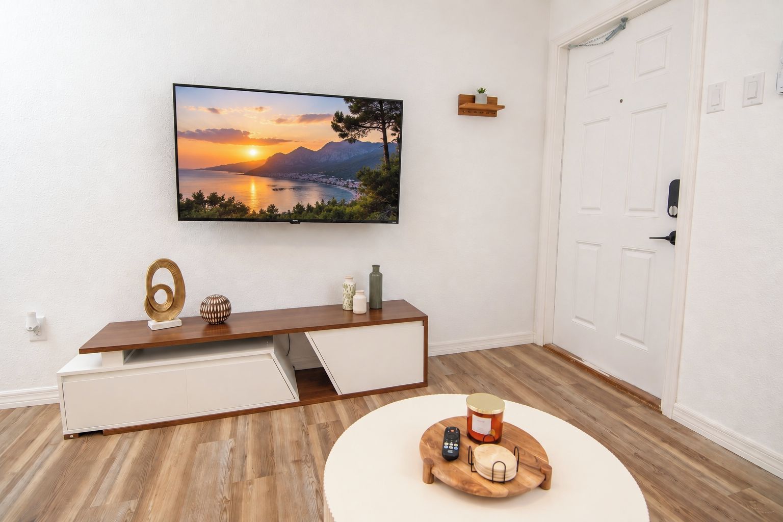A modern living room with a flat-screen TV mounted on the wall, a wooden console table with storage, a round coffee table with a tray, and a white door.