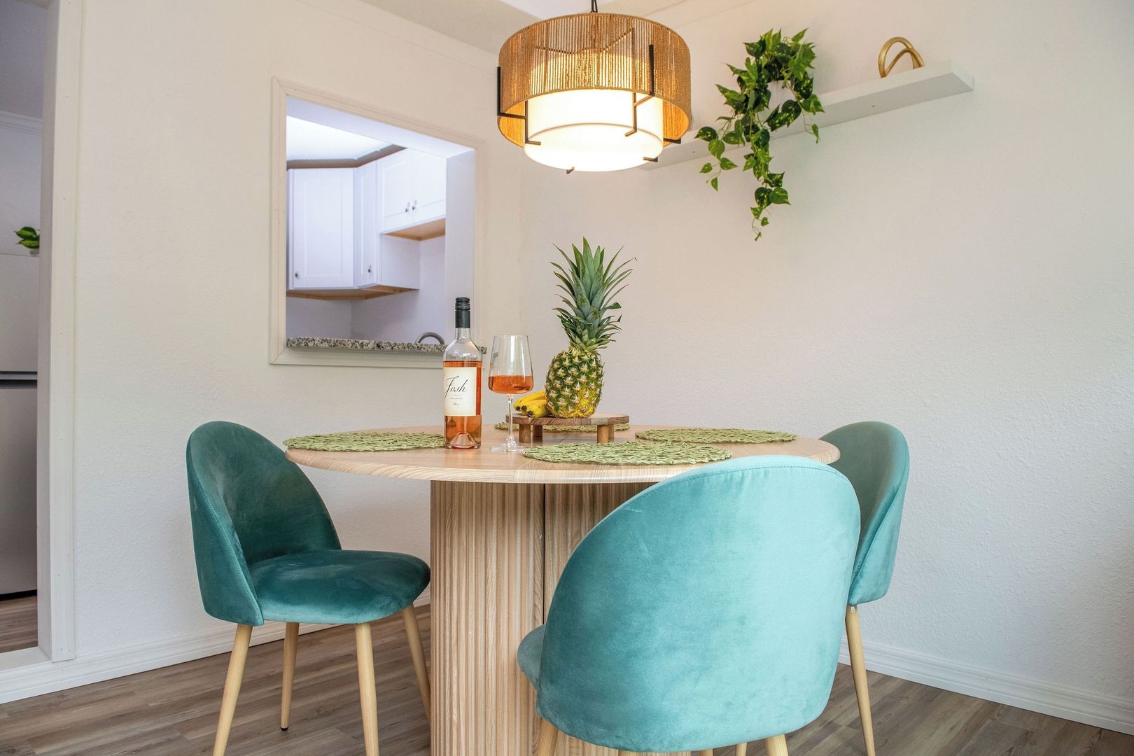 Ein Essbereich mit einem runden Tisch, zwei türkisfarbenen Stühlen, einer Weinflasche, einem Glas und einer Ananas auf dem Tisch.